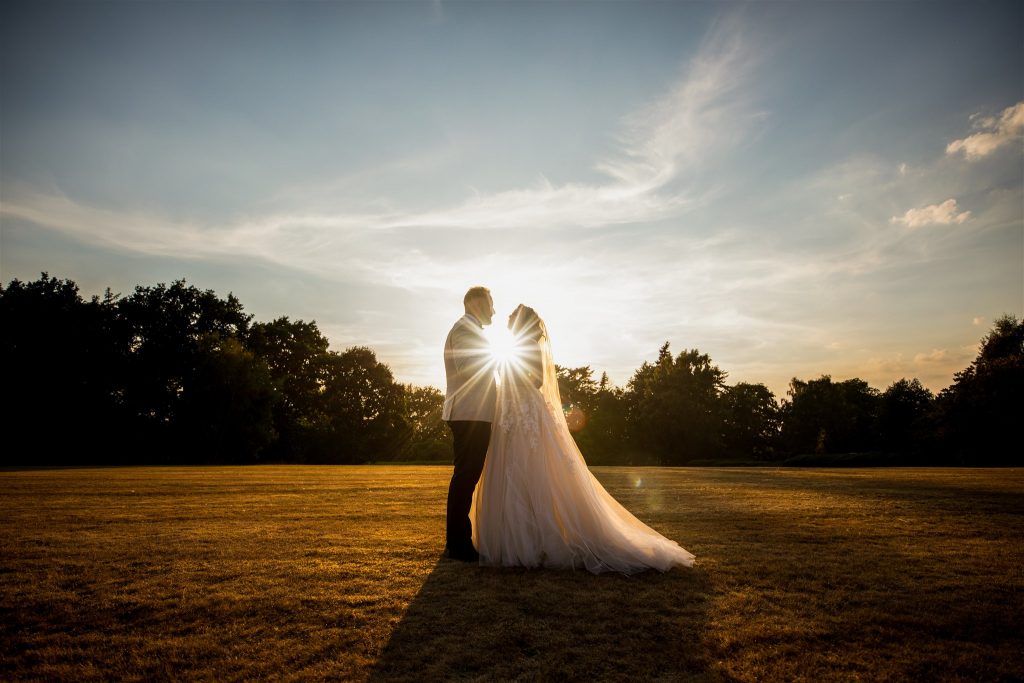 Bride and Groom standing facing each other as the sun sets between them. They are standing in a field with trees in the distance
