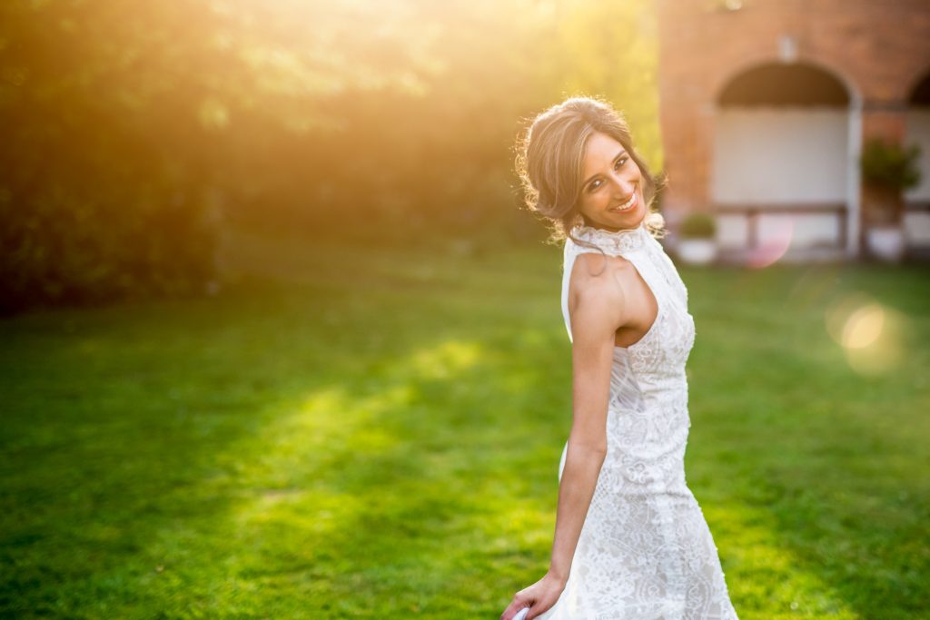 Bride smiling art the camera as she stands on some grass with the sun setting behind her
