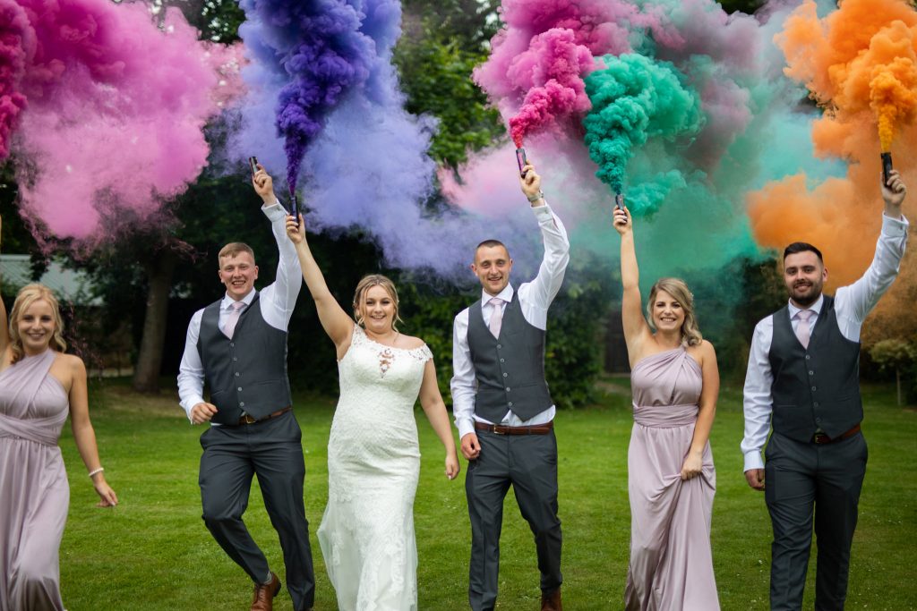 Bride and groom with their friends walking towards a wedding photographer holding up colour smoke flares.